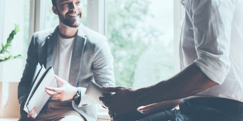 Cropped image of two young businessmen in smart casual wear talking and smiling while sitting on the window sill in office